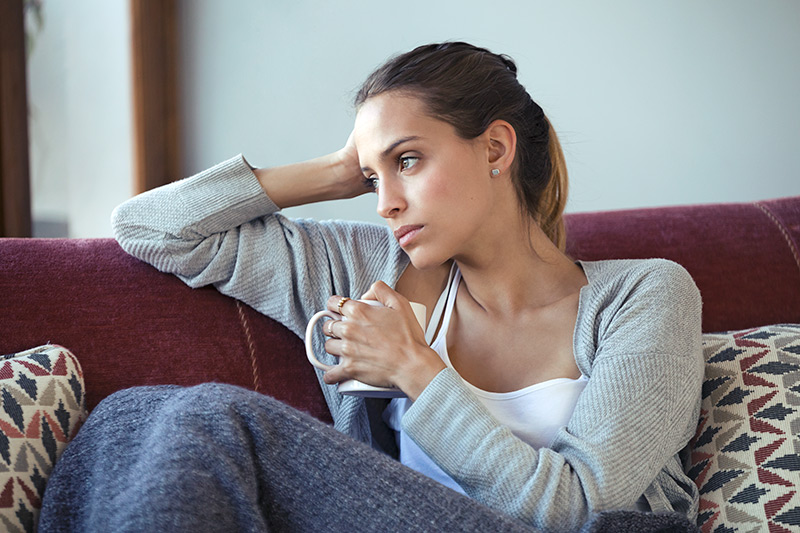 Sad depressed young woman thinking about her problems while drinking coffee on sofa at home