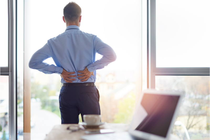 business man standing by his desk looking out a window and holding his back