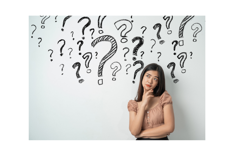 Girl standing with her hand on her chin in front of a bunch of question marks on the wall behind her.