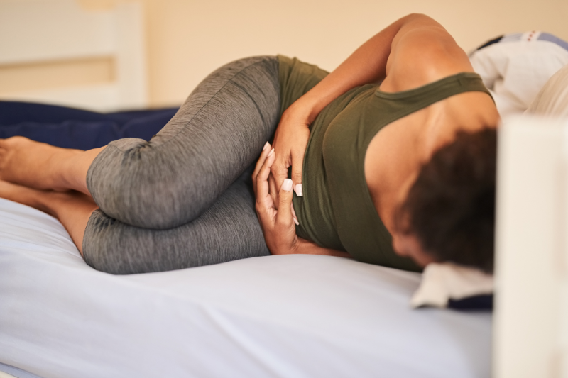 woman in green jumpsuit curled up on bed clutching stomach in pain