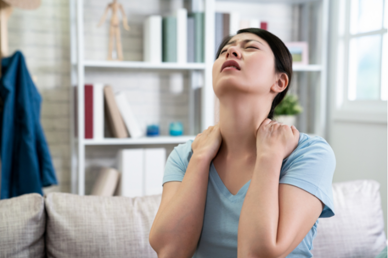woman sitting on couch with arms behind head holding neck in pain